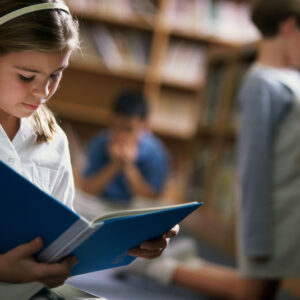 Portrait of young girl reading in library with others kids behind