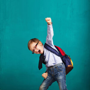 Cheerful smiling little boy with big backpack jumping and having fun against blue wall